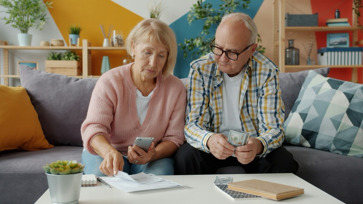 Ein älteres Paar sitzt auf dem Sofa und rechnet mit Rechnungen und Handy.